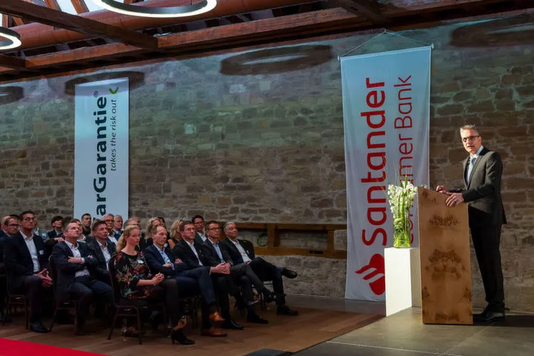 Ein Mann hält eine Rede vor einem sitzenden Publikum in einem Raum mit Bannern der Santander Consumer Bank und CarGarantie.