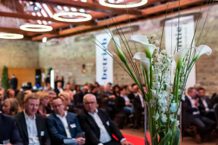 Eine Veranstaltung mit Menschen in einem Raum, im Vordergrund steht eine Vase mit weißen Blumen.
