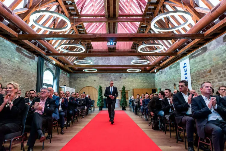 Ein Mann im Anzug geht einen roten Teppich entlang, während ein Publikum in einem Saal applaudiert.