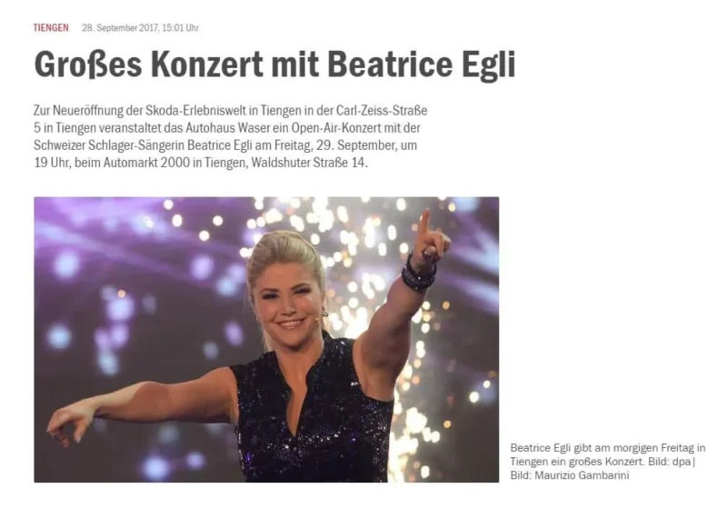 Eine Sängerin in schwarzem Outfit lächelt und hebt einen Arm vor einem funkelnden Hintergrund.