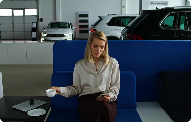 AU UND HAUPTUNTERSUCHUNG Eine Frau sitzt mit einer Tasse in der Hand auf einem Sofa in einem Autohaus, im Hintergrund stehen mehrere Autos.