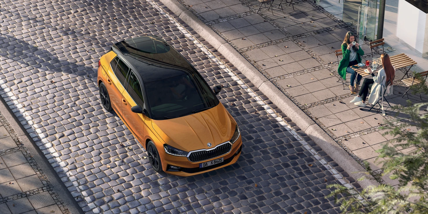 Ein orangefarbener Škoda Fabia fährt auf einer gepflasterten Straße, während zwei Personen in einem Café sitzen.