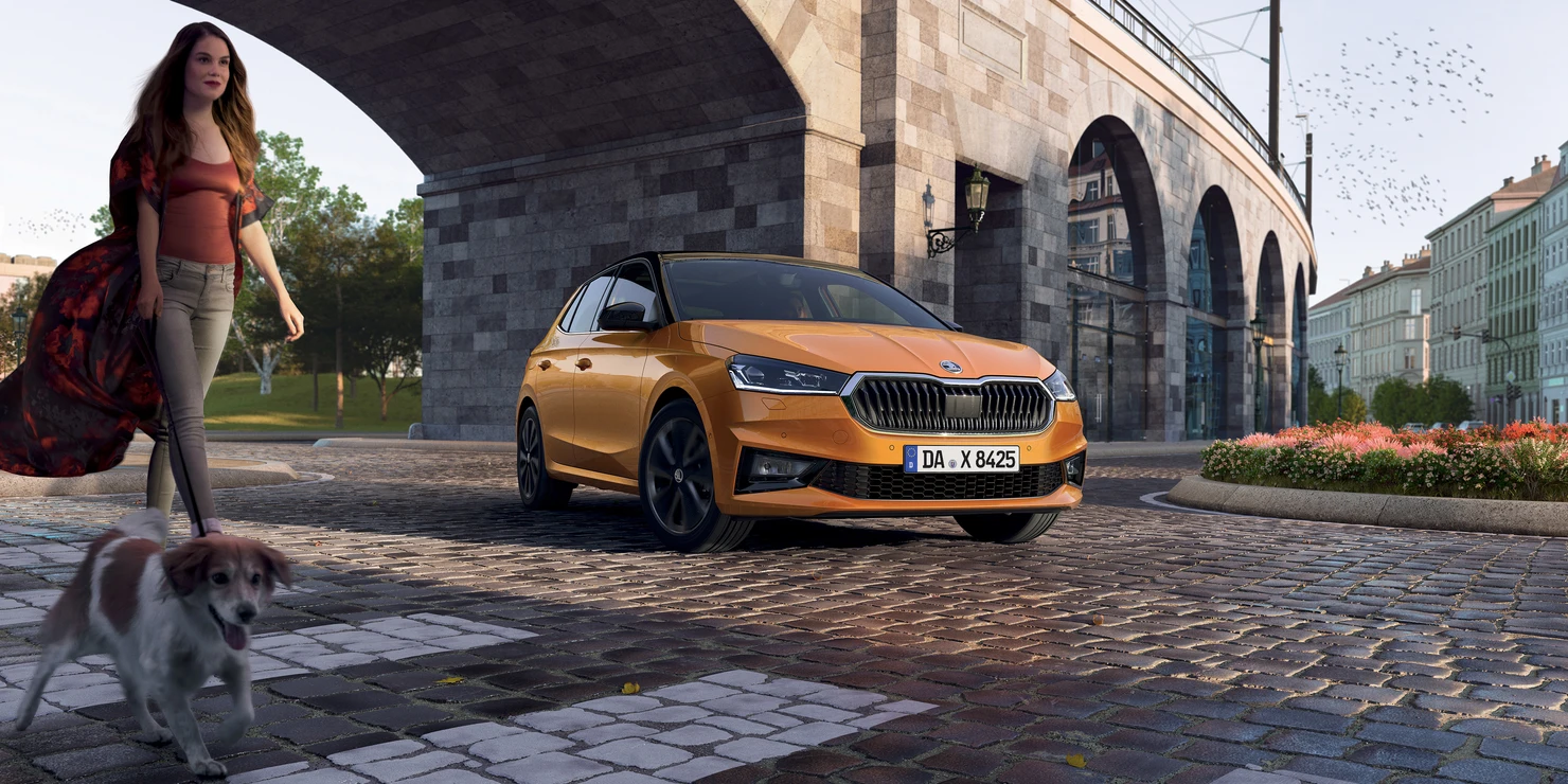 Ein orangefarbener Škoda Fabia parkt auf einer gepflasterten Straße neben einer Frau mit Hund.