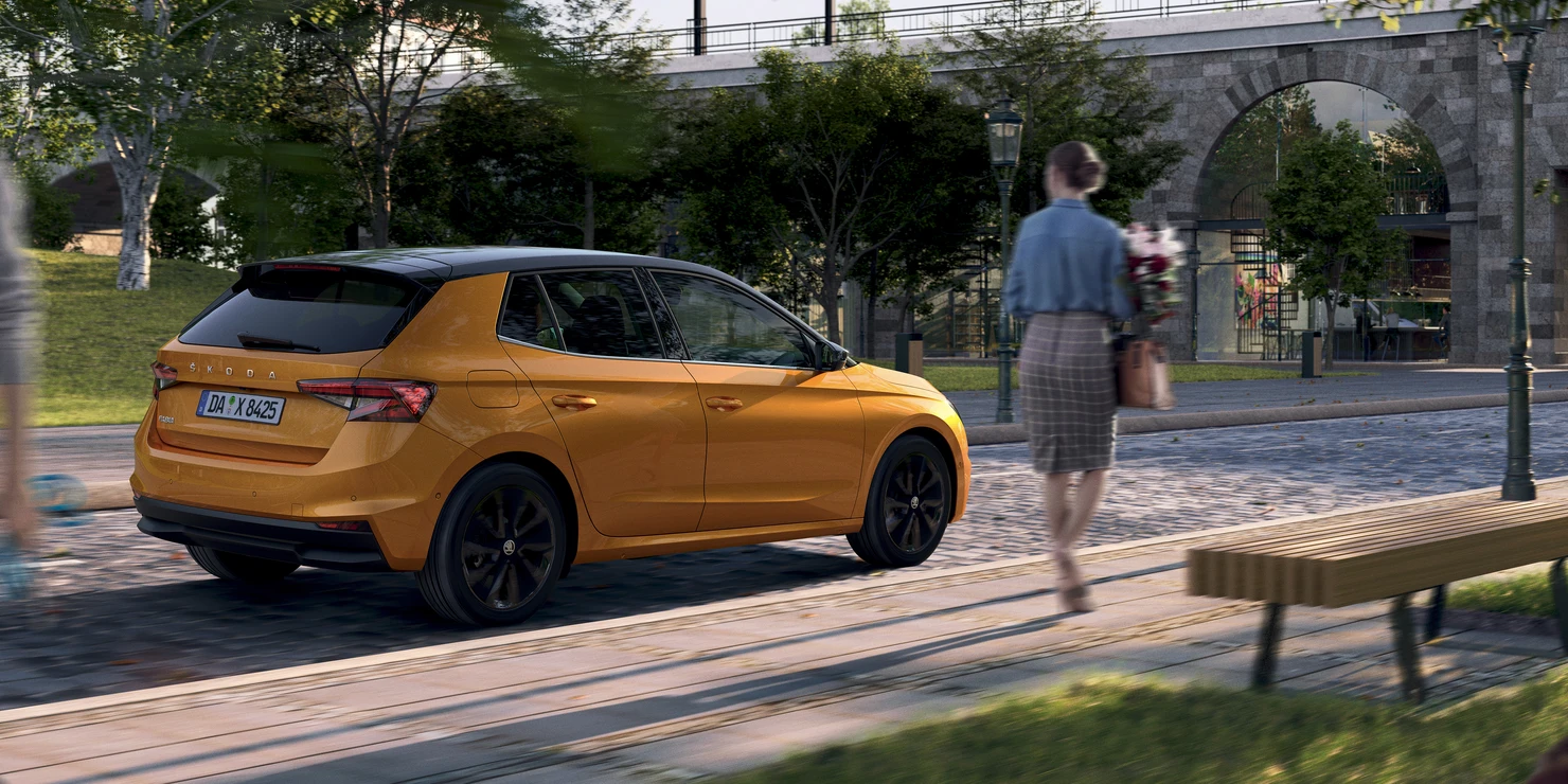 Ein orangefarbener Škoda Fabia parkt auf einer Straße, während eine Person mit Blumen vorbeigeht.