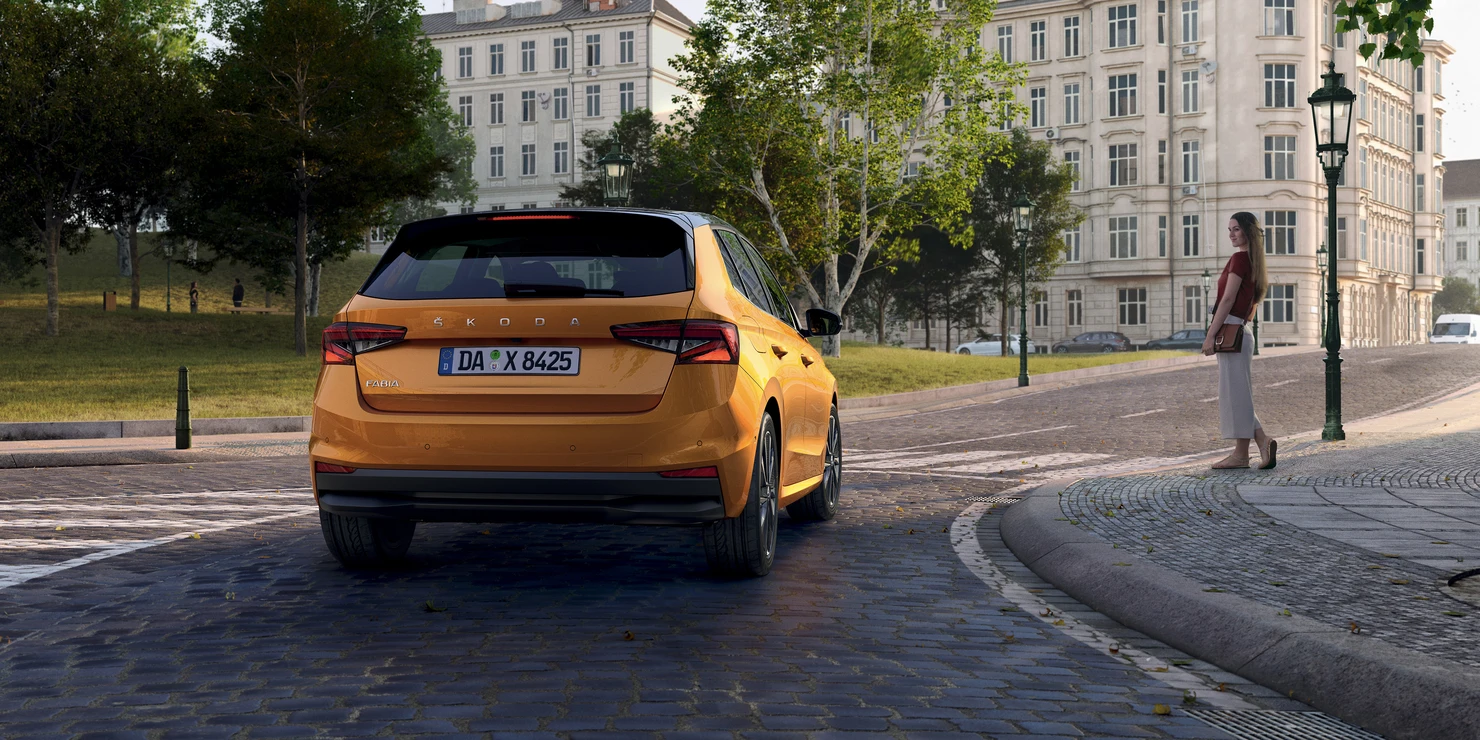 Ein orangefarbener Škoda Fabia parkt auf einer gepflasterten Straße neben einer Frau.