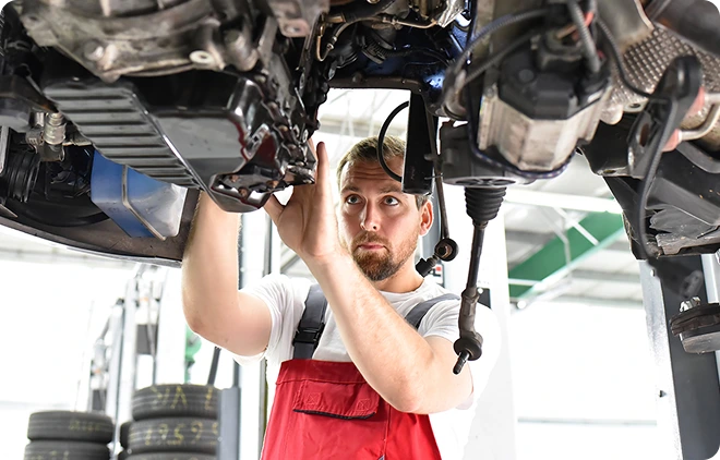 WERKSTATTSERVICE Ein Mechaniker in roter Latzhose arbeitet unter einem Auto in einer Werkstatt.