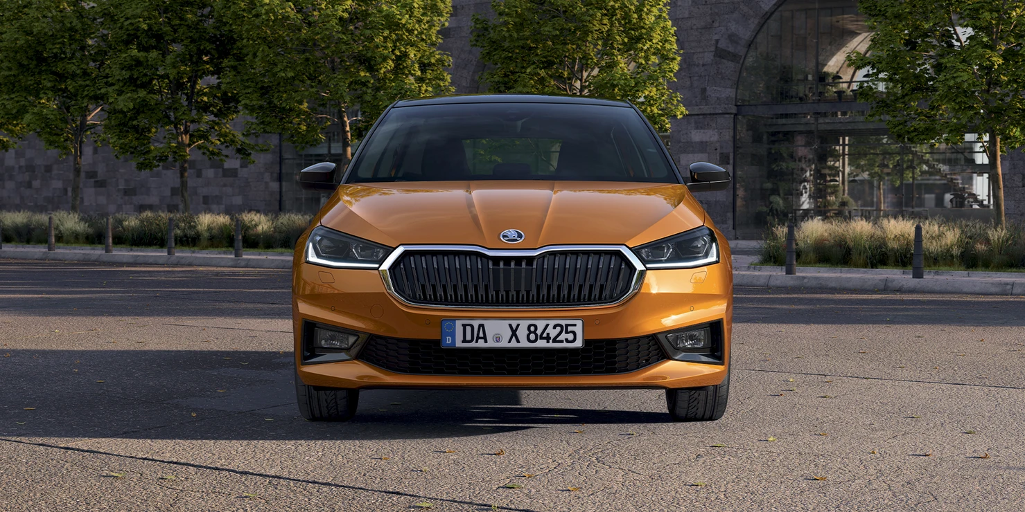 Ein orangefarbener Skoda Fabia steht auf einer Straße vor einer Steinmauer und Bäumen.