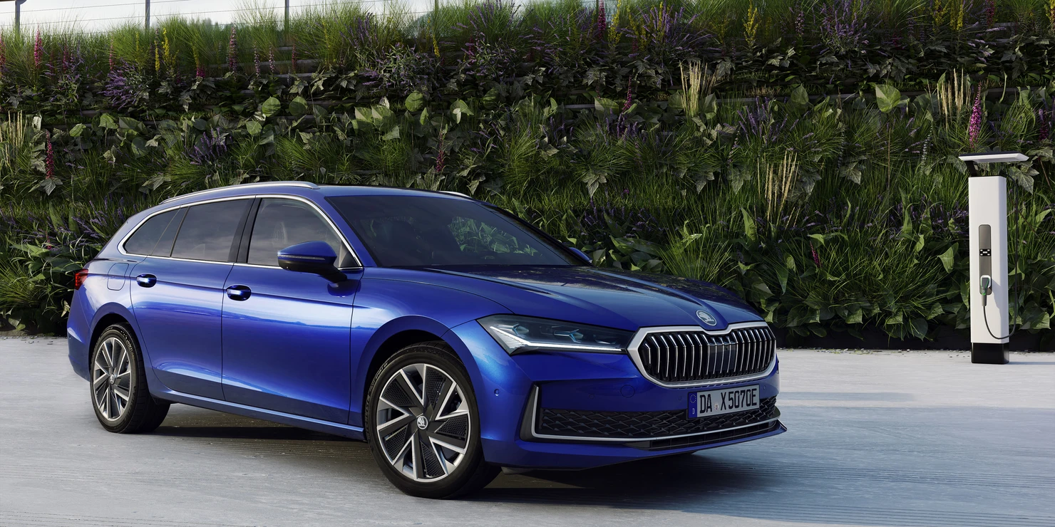 Ein blauer Škoda Superb Combi iV steht neben einer Ladestation vor einer grünen Wand.