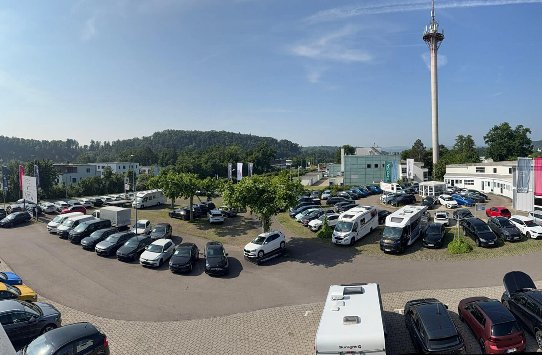 Ein großer Parkplatz mit verschiedenen Autos und Wohnmobilen vor einem Autohaus mit einem hohen Sendeturm im Hintergrund.