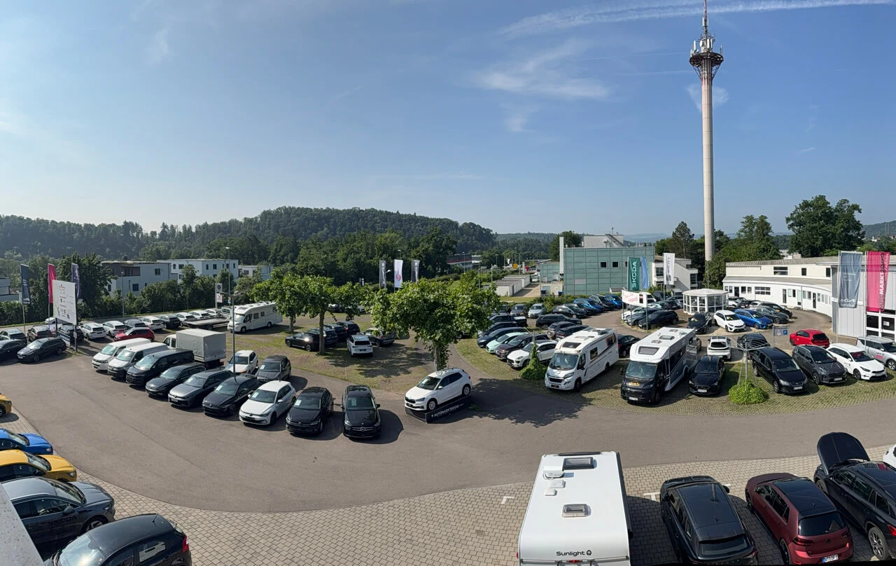 Groß-_IMG_9385__1 Ein Autohaus mit einer Vielzahl geparkter Fahrzeuge, darunter Autos und Wohnmobile, sowie einem hohen Sendeturm im Hintergrund.