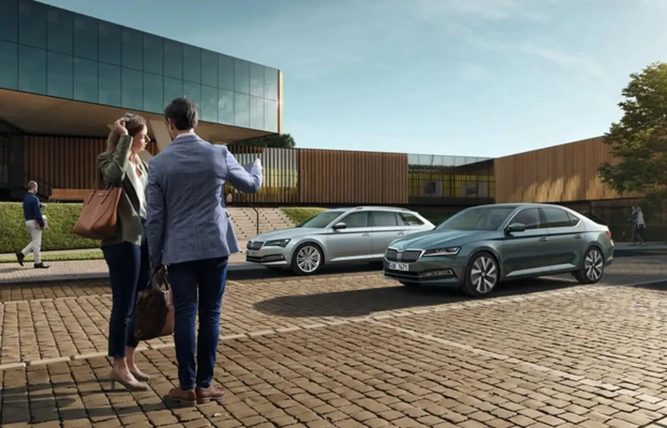 mobilitaetsgarantie-p1920_1 Zwei Personen stehen vor einem modernen Gebäude mit geparkten Skoda Superb Autos.