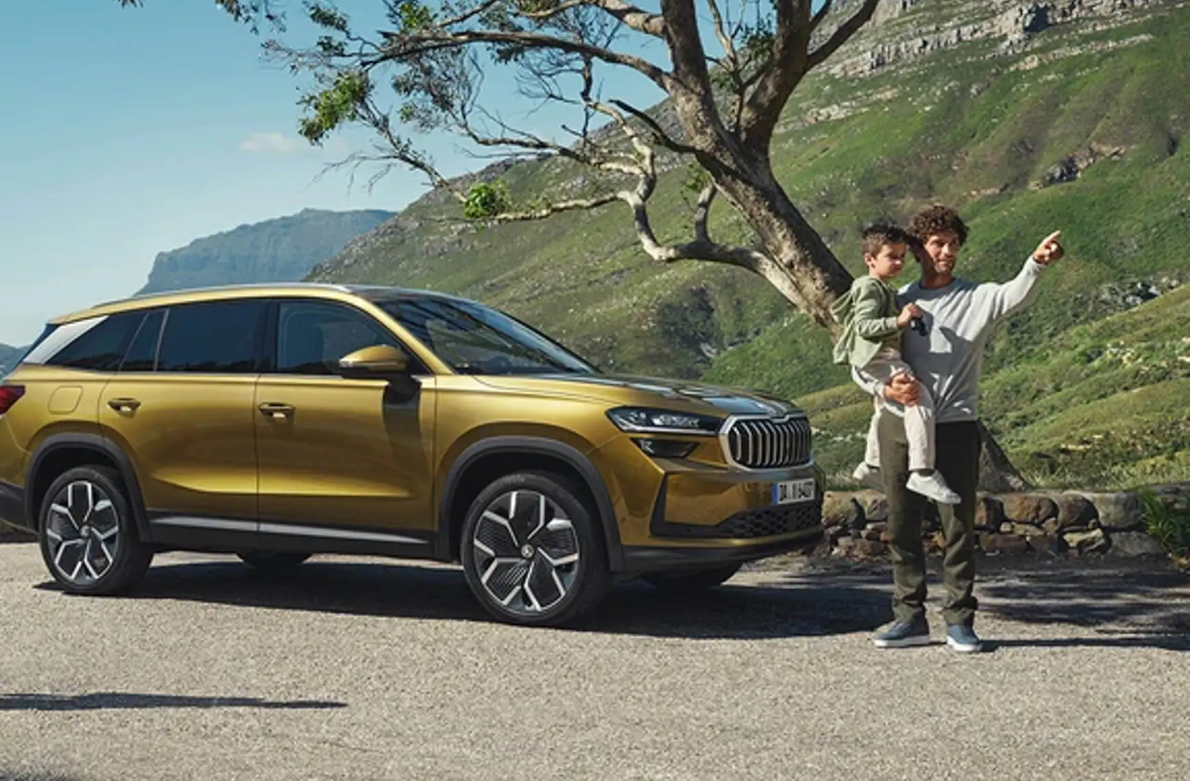 Ein Mann hält ein Kind und zeigt auf etwas neben einem goldenen Skoda-SUV vor einer grünen Landschaft.