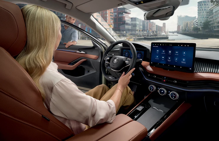 superb-combi-gg-04 Eine Frau sitzt am Steuer eines Škoda Superb Combi mit einem großen Touchscreen-Display, während eine Person von außen in das Auto schaut.