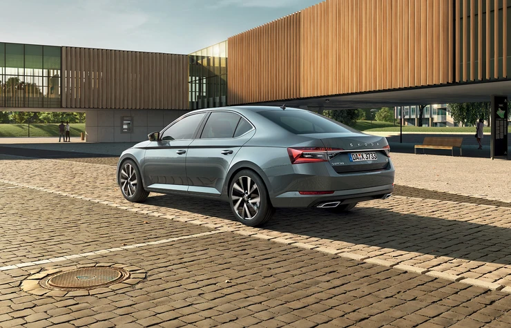 superb-gg-01 Ein grauer Škoda Superb steht auf einem gepflasterten Platz vor einem modernen Gebäude.