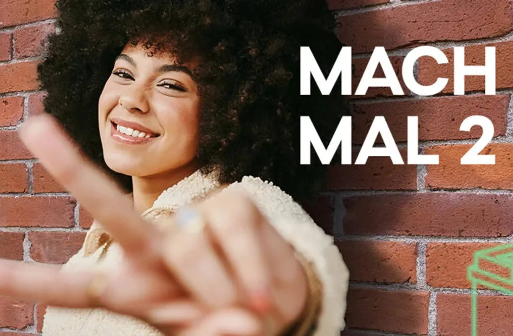 Eine lächelnde Person zeigt ein Peace-Zeichen vor einer Backsteinmauer, daneben der Text "Mach mal 2".