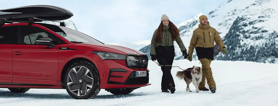 AutohausWaser_Service Winter Menschen spazieren mit einem Hund neben einem roten Skoda Enyaq im Schnee.